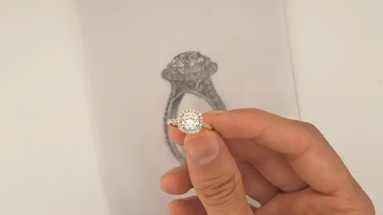 A jewelry designer meticulously sketches a detailed ring design in a studio, with crafting tools and materials visible in the background.