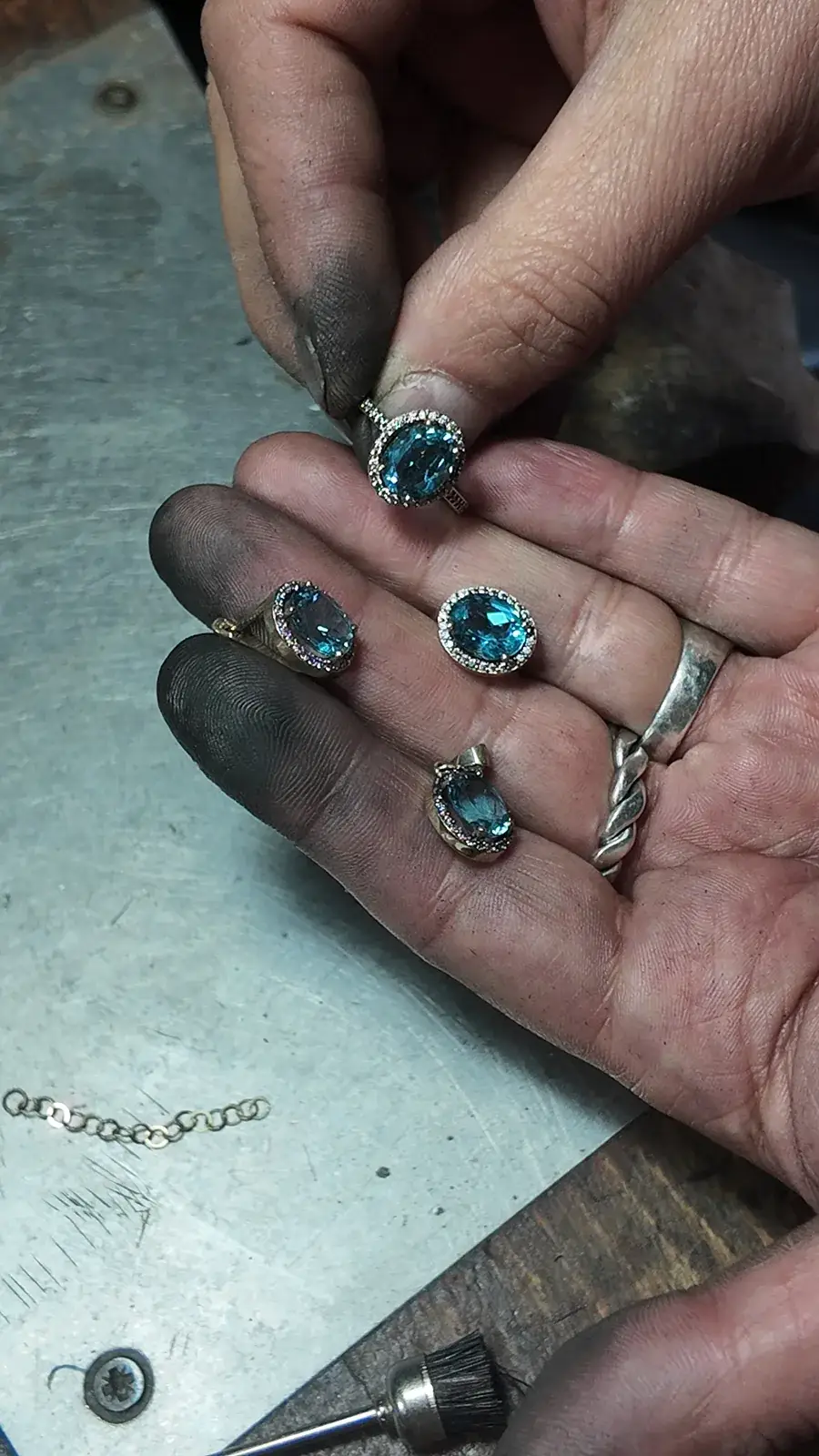 Hands holding jewelry pieces with blue-green stones above a jeweler’s bench